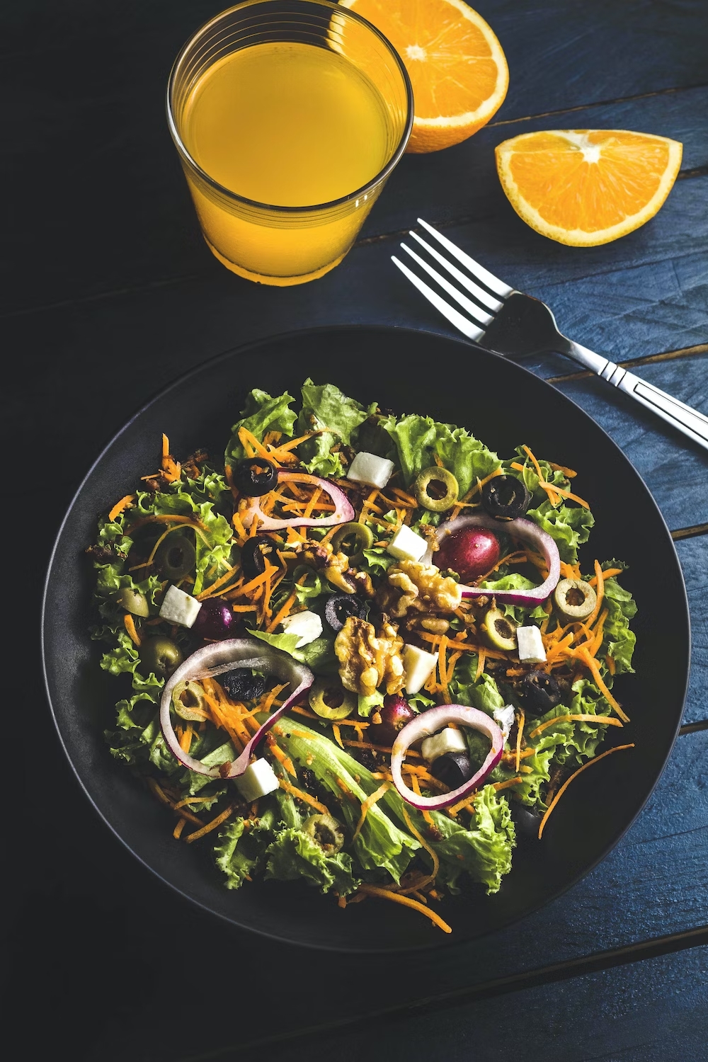 A Salad with lettuce, onions, olives, shredded carrot. A fork is on the side as well as a glass of orange juice with sliced oranges next to it.